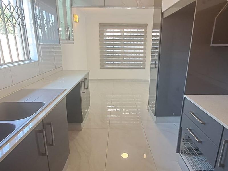 A modern, clean kitchen featuring sleek cabinetry, stone countertops, a double basin sink, and polished tiled flooring, leading into an open-plan living area.