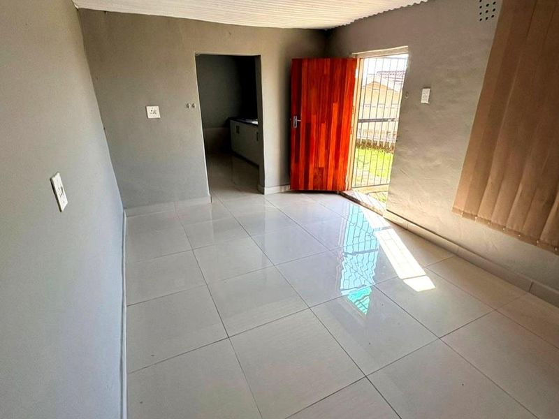 An empty living area featuring tiled flooring, a timber door with a security gate, and access to an adjacent kitchen space.