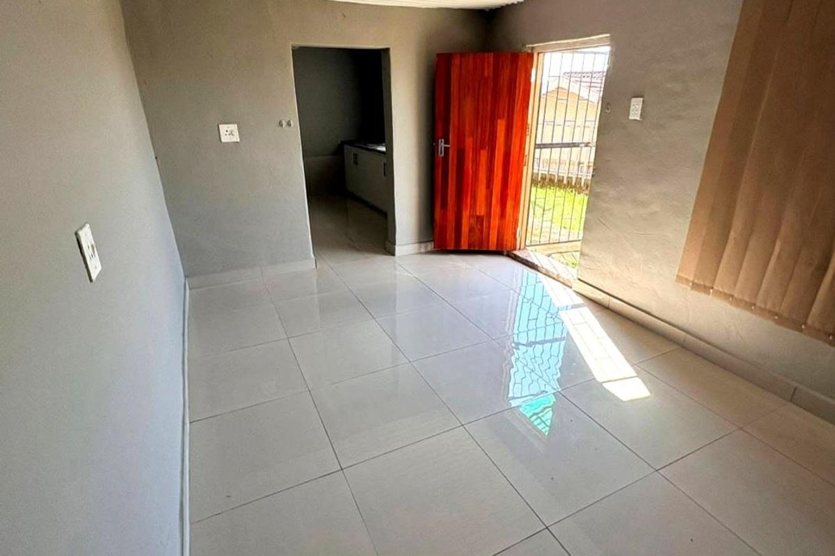 An empty living area featuring tiled flooring, a timber door with a security gate, and access to an adjacent kitchen space.