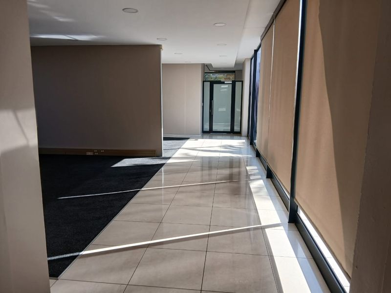 A bright, open-plan office space featuring a mix of tiled and carpeted flooring, large windows with roller blinds, and recessed ceiling lighting.