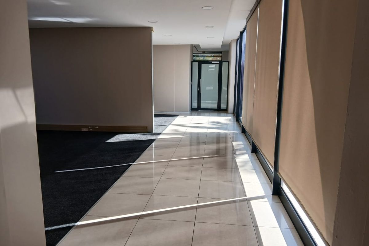A bright, open-plan office space featuring a mix of tiled and carpeted flooring, large windows with roller blinds, and recessed ceiling lighting.