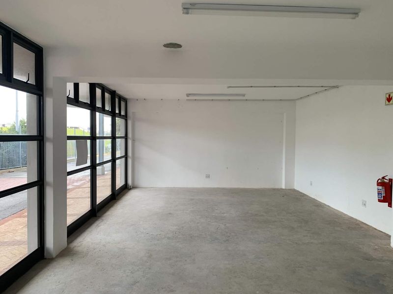 An empty, open-plan commercial space featuring large black-framed glass windows, concrete flooring, and functional overhead lighting, suitable for various business uses.