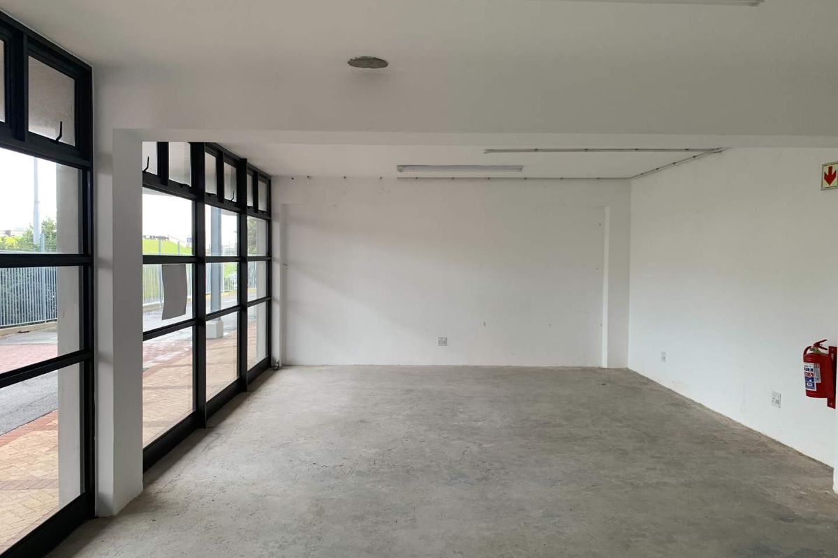 An empty, open-plan commercial space featuring large black-framed glass windows, concrete flooring, and functional overhead lighting, suitable for various business uses.