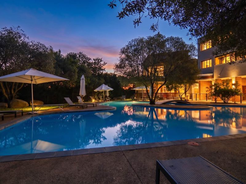 A beautifully lit swimming pool area at dusk, featuring paved surrounds, mature trees, and direct access to the apartment building exterior.