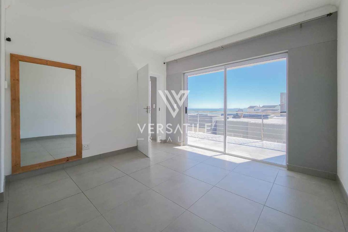 A bright, empty bedroom featuring large sliding glass doors that open onto a balcony with ocean views and neutral tiled flooring.