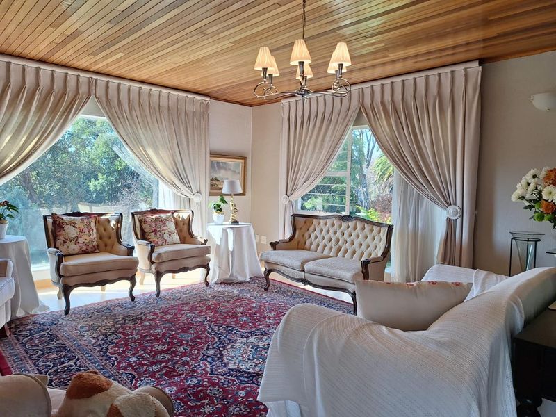 A bright and spacious living room featuring a warm timber-clad ceiling, large windows with natural light, and a classic chandelier.