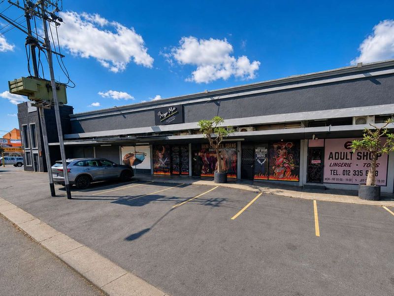 Front exterior view of a retail property featuring a paved parking area, shopfront glazing, and a concrete facade with signage.