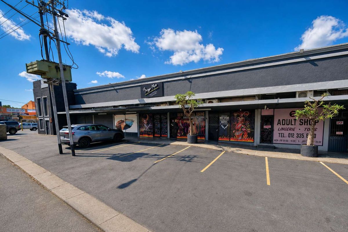 Front exterior view of a retail property featuring a paved parking area, shopfront glazing, and a concrete facade with signage.