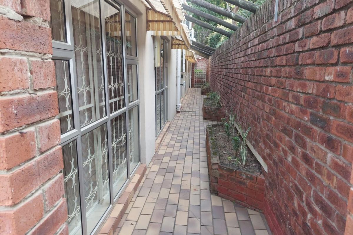A narrow paved side walkway featuring built-in brick planters, a timber pergola structure, and secure aluminium-framed windows along the exterior wall.