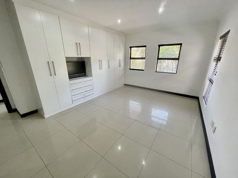 A bright, modern bedroom featuring extensive white built-in cabinetry, polished tiled flooring, and large windows with fitted venetian blinds.