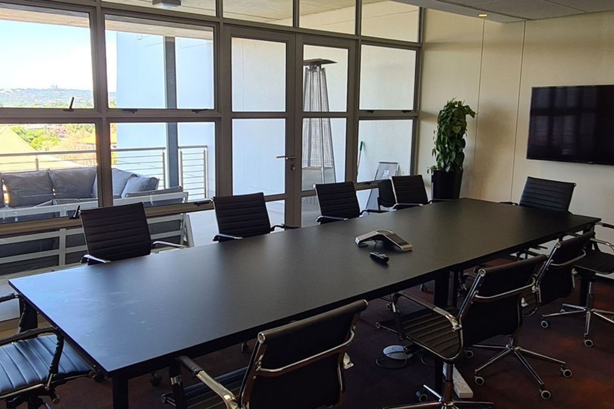 A professional boardroom featuring a large conference table, wall-mounted screen, and floor-to-ceiling glass partitions providing access to an outdoor balcony area.