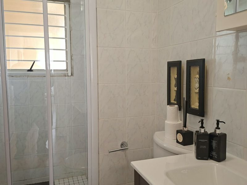 A functional bathroom featuring a shower enclosure, vanity basin, and toilet, finished with neutral wall tiles and security bars on the window.