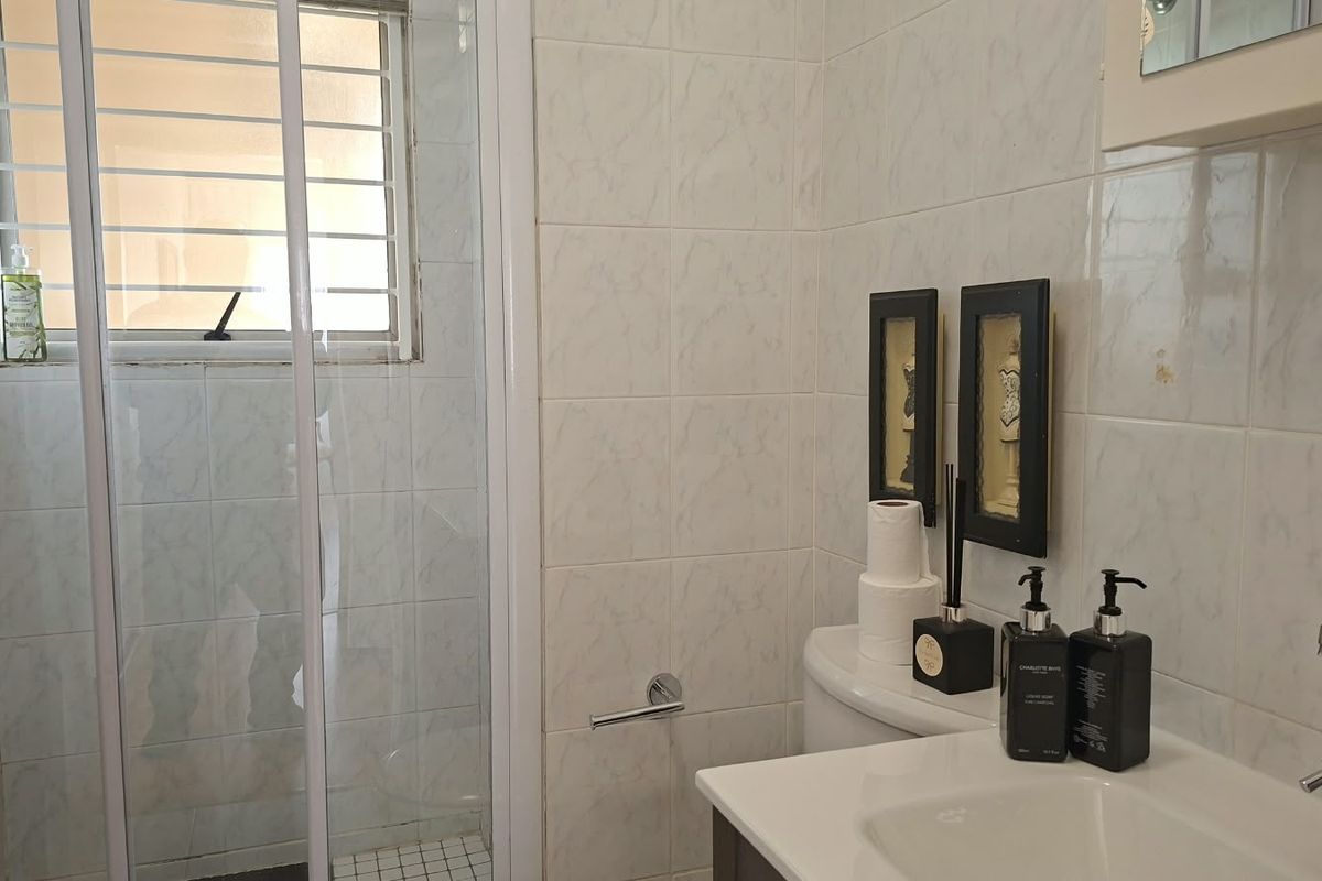 A functional bathroom featuring a shower enclosure, vanity basin, and toilet, finished with neutral wall tiles and security bars on the window.