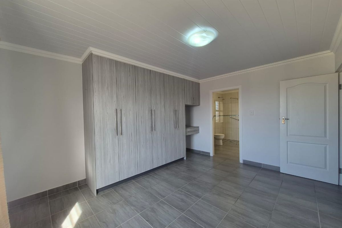 A clean, empty bedroom featuring modern grey wood-look built-in cupboards, tiled flooring, and a tongue and groove ceiling with direct access to an en-suite bathroom.