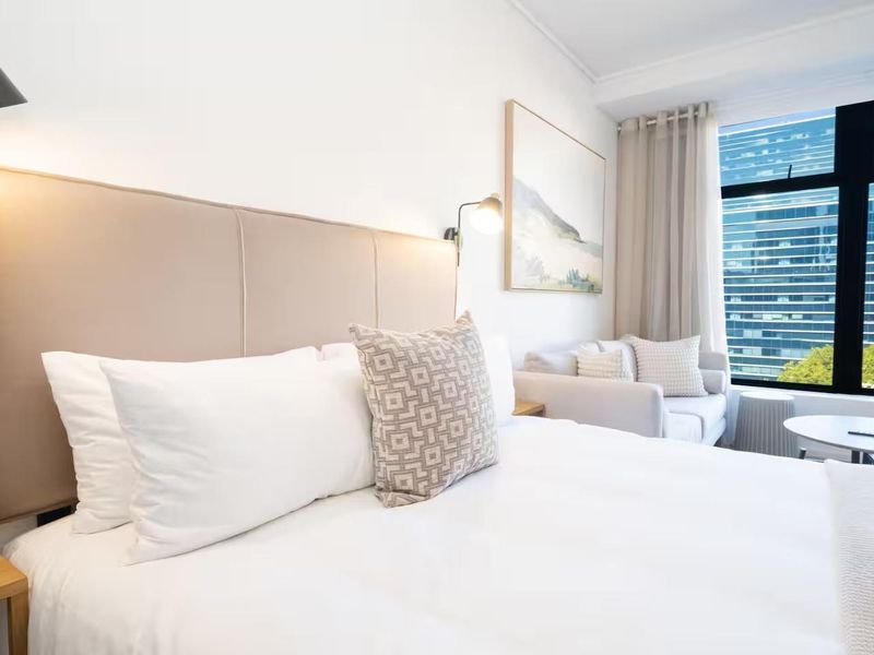 A well-lit, modern bedroom featuring a large window with city views, a built-in headboard, and integrated wall-mounted lighting.