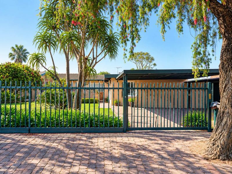 Front view of the property featuring a secure metal fence, paved driveway, carport, and a well-maintained garden area.