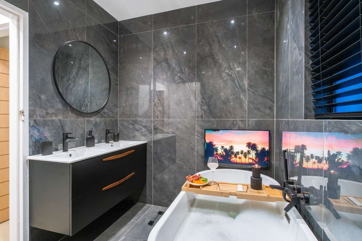 Modern bathroom featuring dark marble-effect wall tiles, a sleek double vanity, and a freestanding bathtub with an integrated wall-mounted television.
