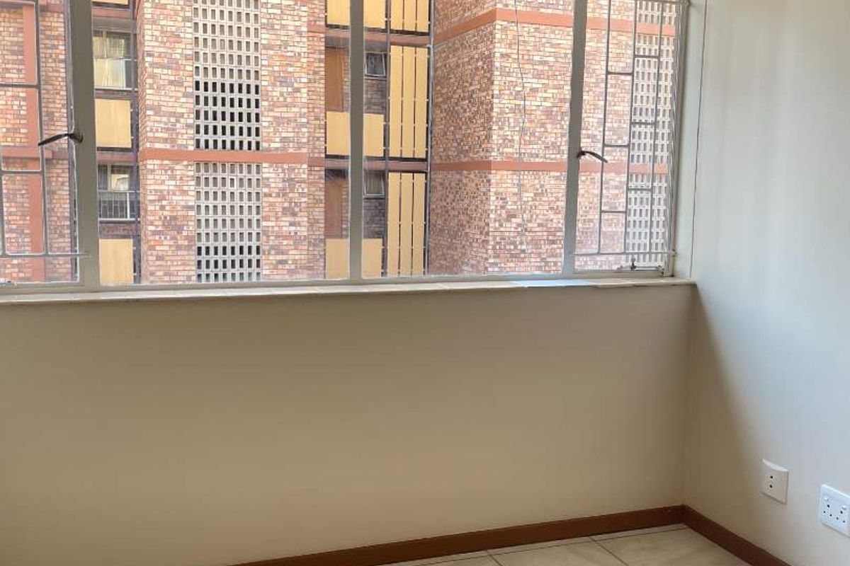 A bright, empty bedroom featuring large windows with an urban view, neutral tiled flooring, and clean wall finishes.