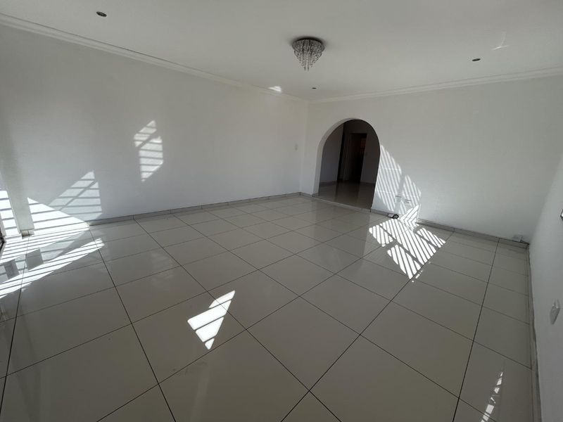 A bright, spacious, and empty living area featuring neutral tiled flooring, recessed lighting, and a decorative archway leading to an adjacent room.