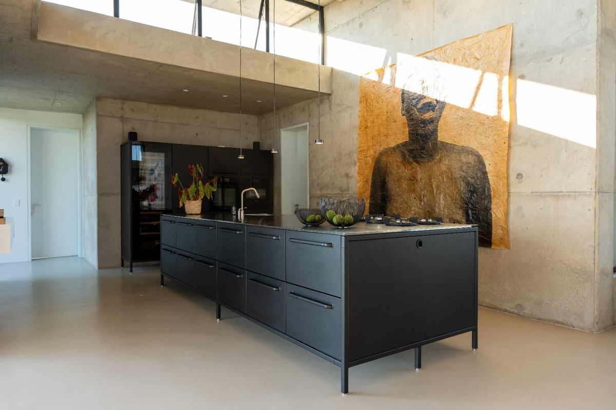 Modern open-plan kitchen featuring a large black central island with integrated cooktop, sleek cabinetry, and striking raw concrete walls under a high ceiling.