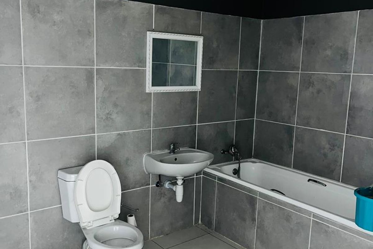 A functional bathroom featuring grey wall tiles, a built-in bathtub, a wall-mounted basin, and a standard toilet.
