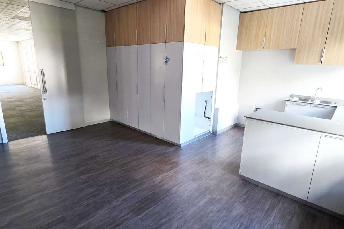 A functional office kitchenette area featuring built-in cabinetry, a sink, and wood-look flooring, with an adjacent office space visible through the doorway.