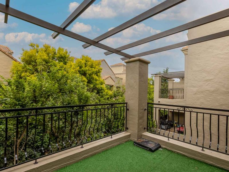 A private balcony featuring a modern pergola structure, wrought iron railings, and artificial turf flooring, overlooking lush greenery.