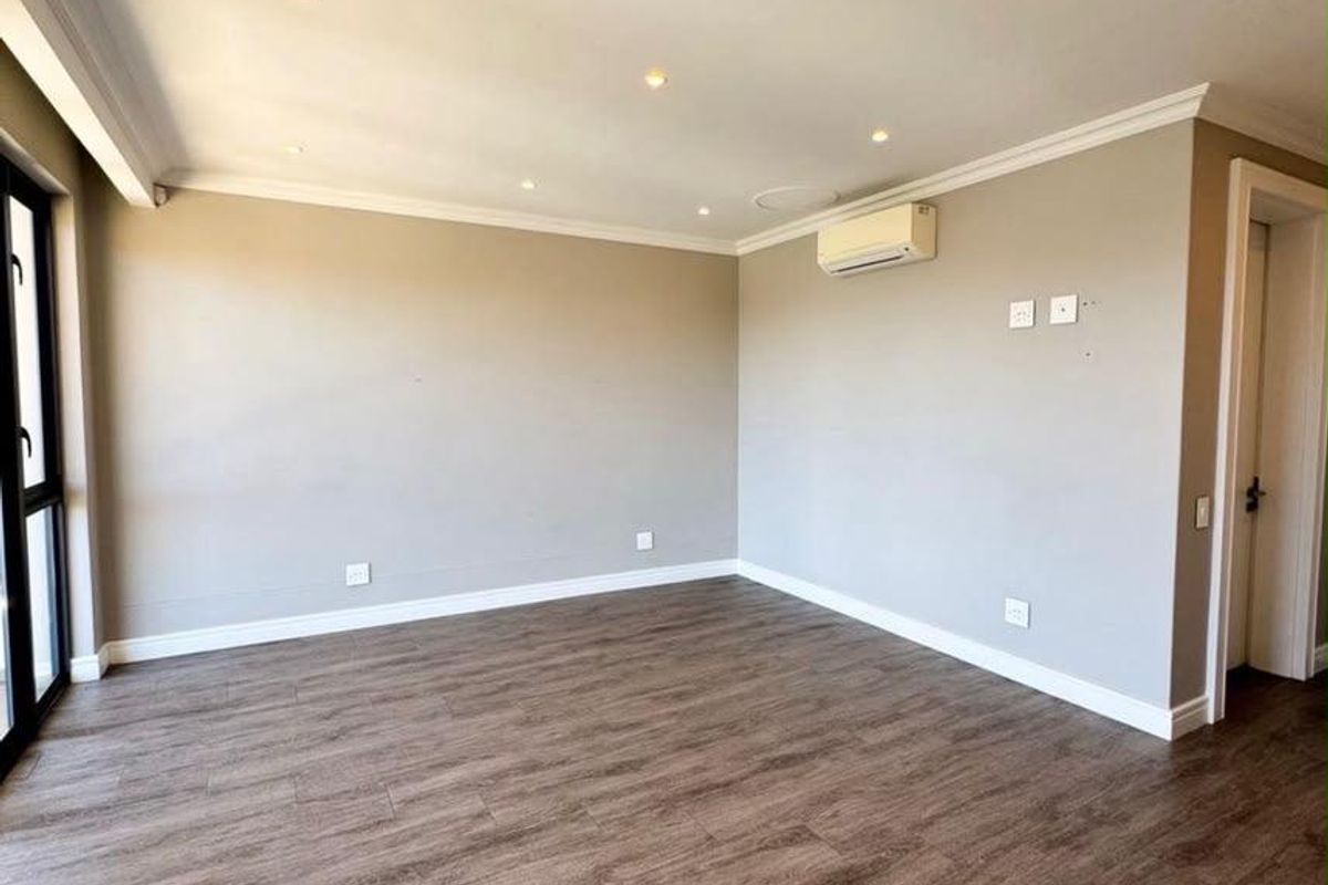 A bright, empty living room featuring modern laminate flooring, recessed downlighting, coving, and a wall-mounted split-system air conditioner.