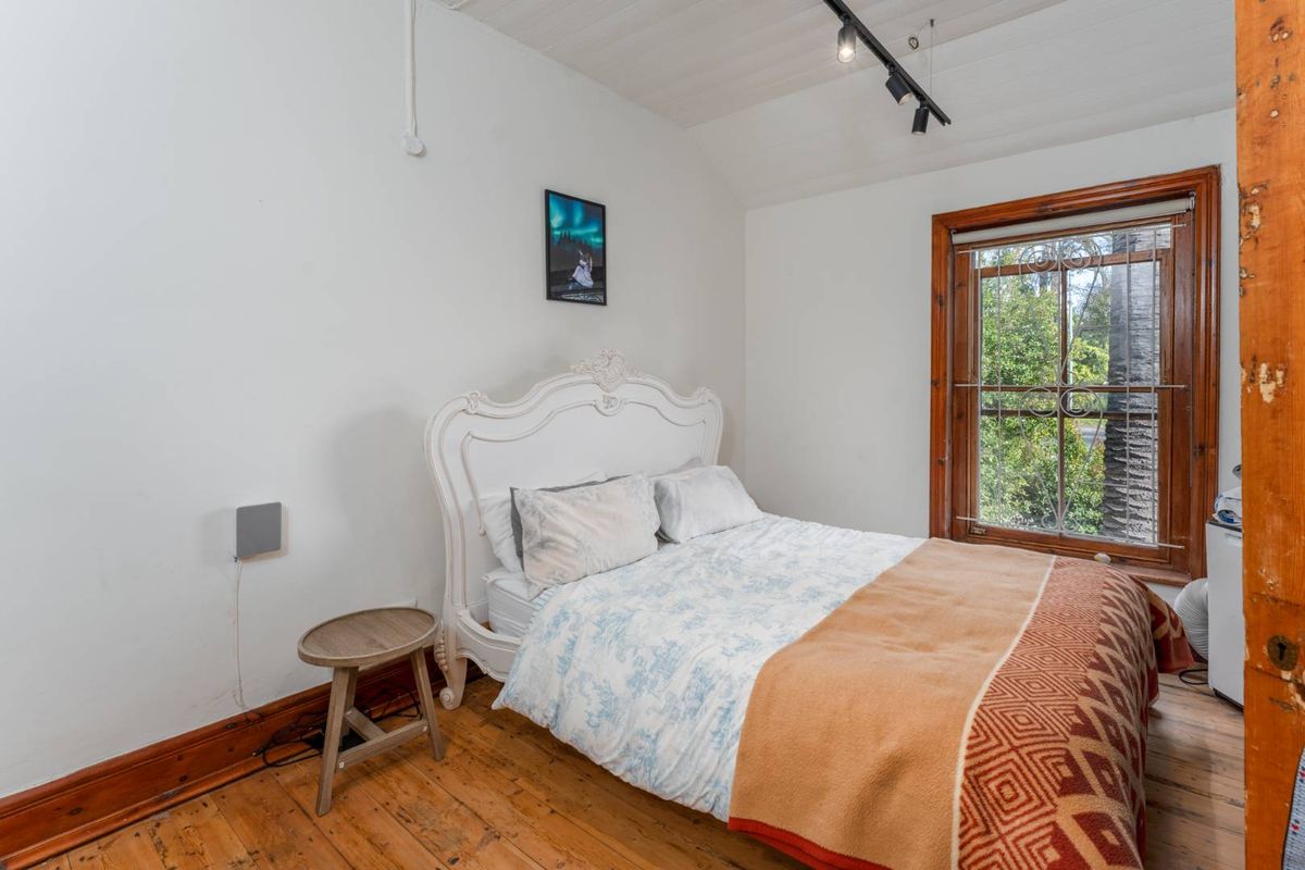 A bedroom featuring timber flooring, track lighting, and a window with security bars, showing signs of wear on the timber finishes.