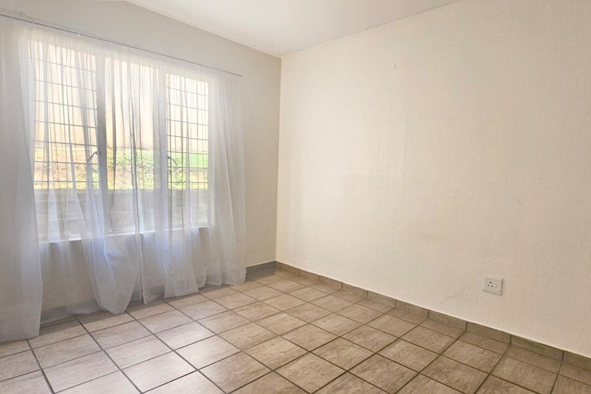 A bright, empty bedroom featuring neutral tiled flooring and a large window with security bars, providing a clean and versatile living space.
