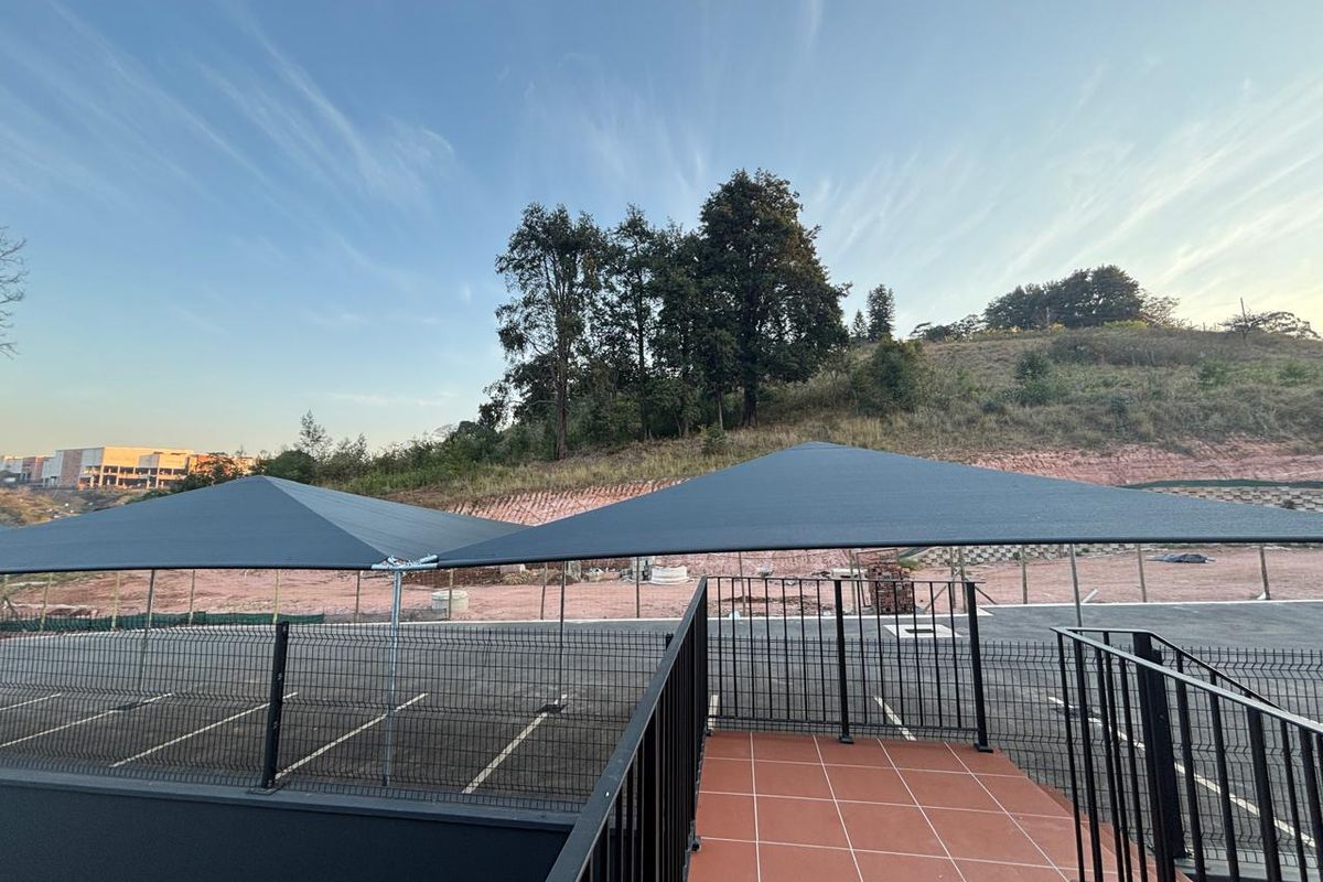 An outdoor area featuring a tiled patio, metal security fencing, and shade sails covering a paved parking space with a hillside backdrop.