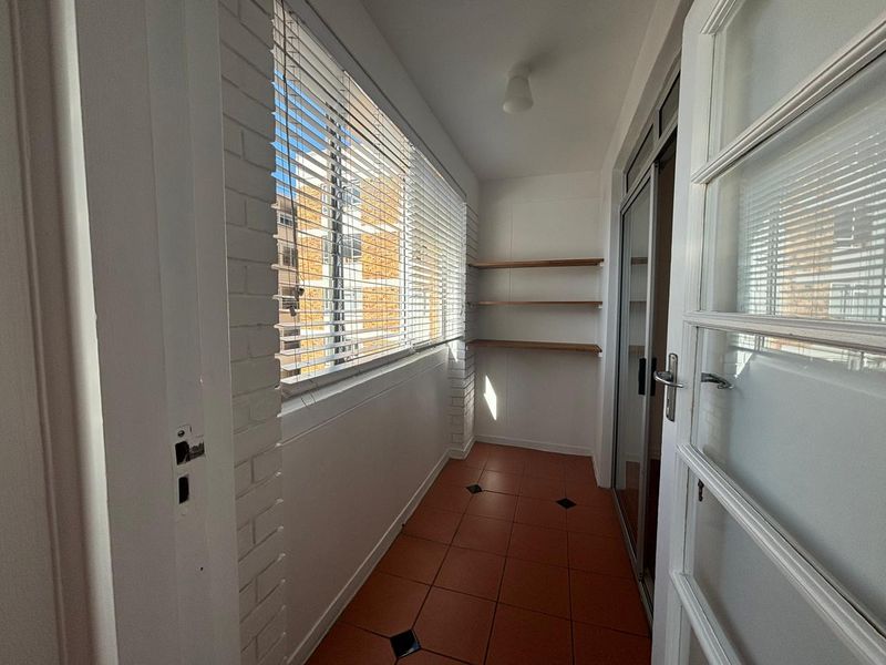 A bright, enclosed balcony featuring tiled flooring, built-in shelving, and large windows with venetian blinds, offering a versatile space for relaxation or storage.