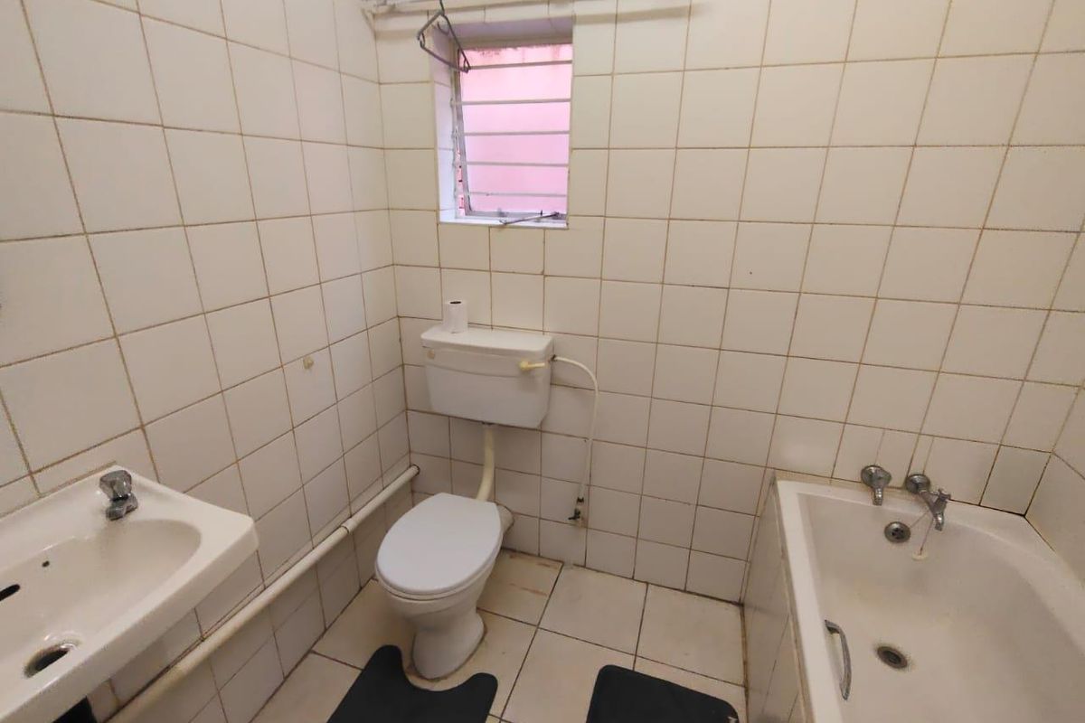 A functional bathroom featuring a standard bathtub, pedestal basin, and toilet with white wall tiling and a small window with security bars.