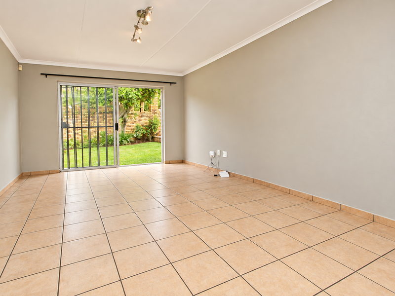 A bright, empty living room featuring neutral walls, ceramic floor tiles, and a sliding glass door providing direct access to the garden.