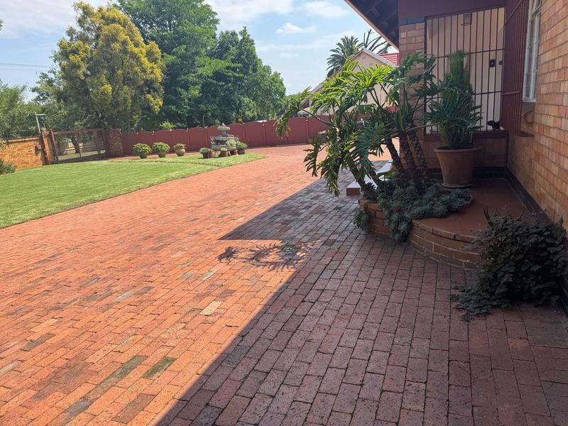 A well-maintained front exterior featuring a spacious brick driveway, landscaped lawn, and secure boundary wall with gated access.