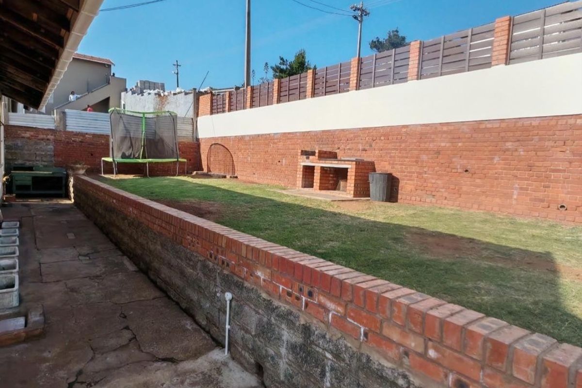 Rear garden area featuring a paved walkway, raised lawn section, and a built-in brick braai against a perimeter wall.