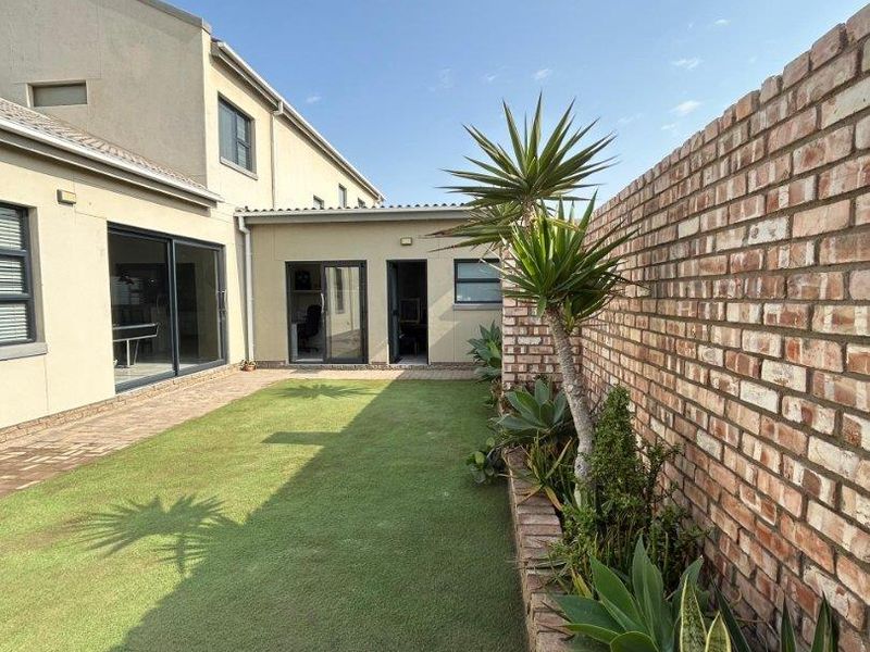 A well-maintained rear garden featuring a neat artificial lawn, paved patio area, and a rustic brick boundary wall with established garden beds.