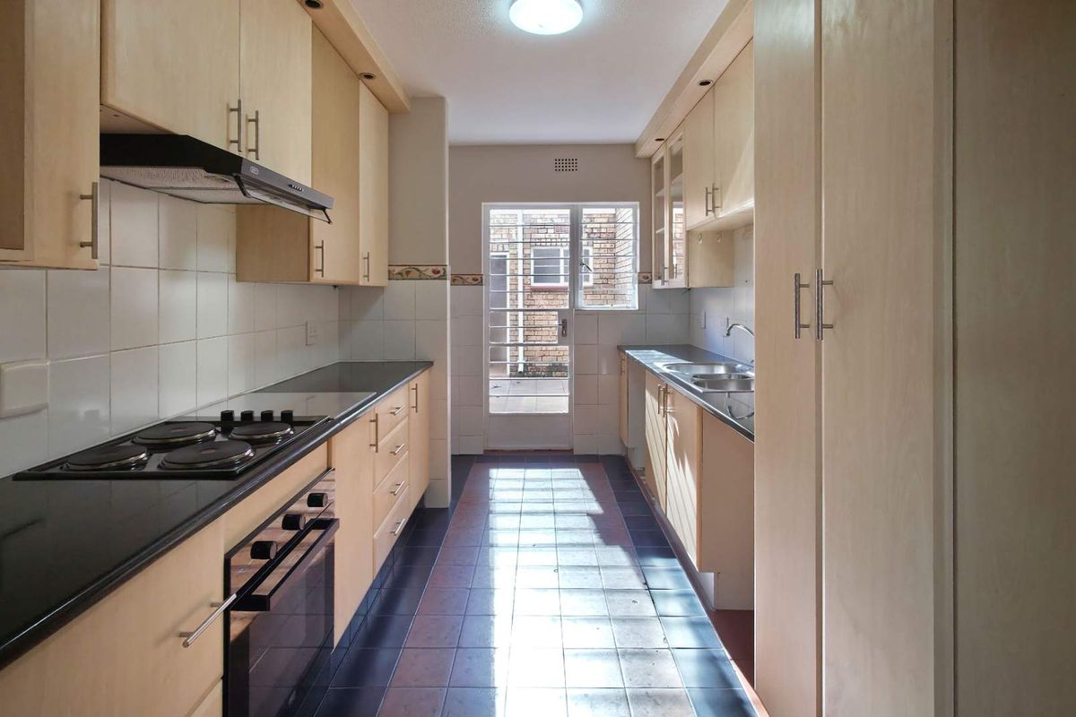 Galley-style kitchen featuring light wood-finish cabinetry, dark countertops, built-in oven, electric hob, extractor fan, and direct access to an exterior courtyard.