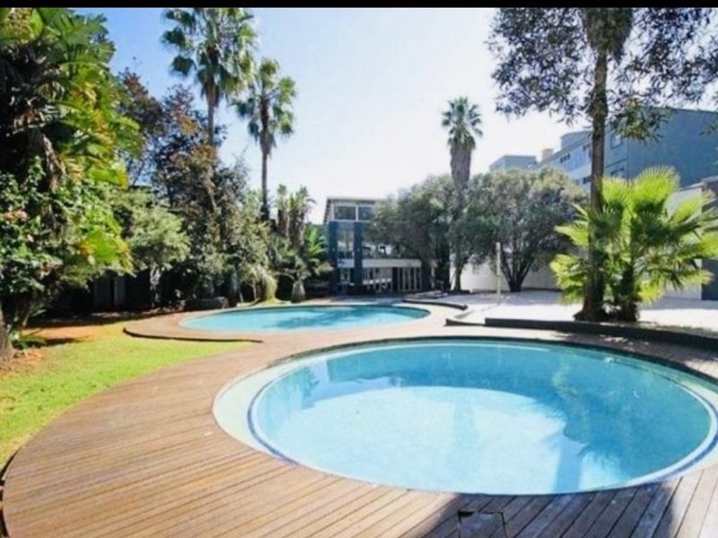 A well-maintained communal pool area featuring circular swimming pools surrounded by timber decking and established landscaped gardens.