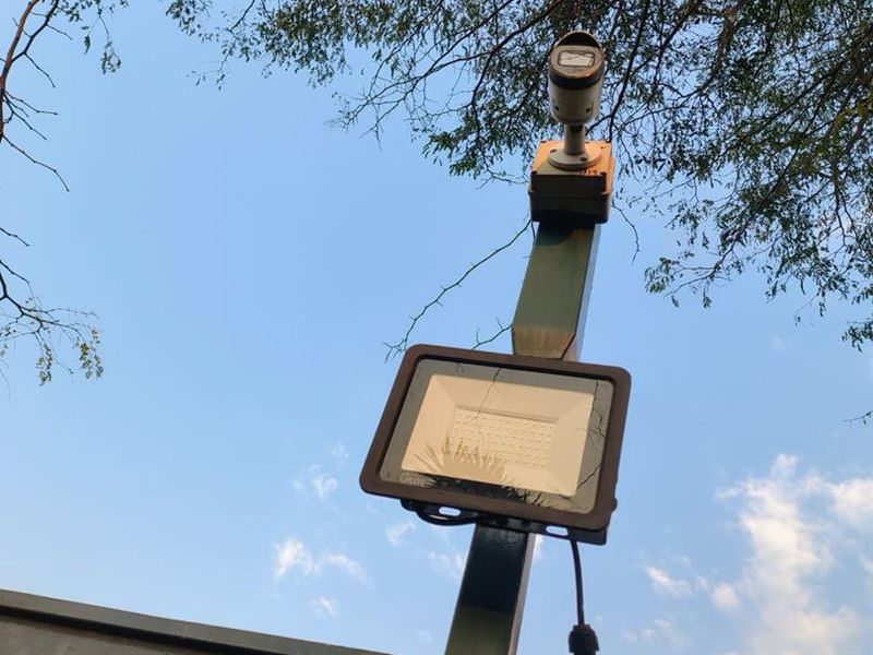 A close-up view of a security camera and LED floodlight mounted on a metal pole against a clear sky.