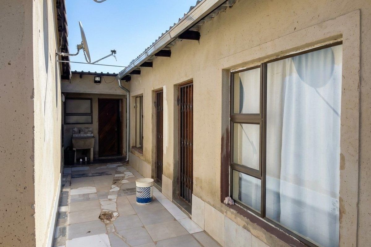 A narrow rear exterior walkway featuring tiled flooring, security gates, and a built-in concrete wash basin along the side of the house.