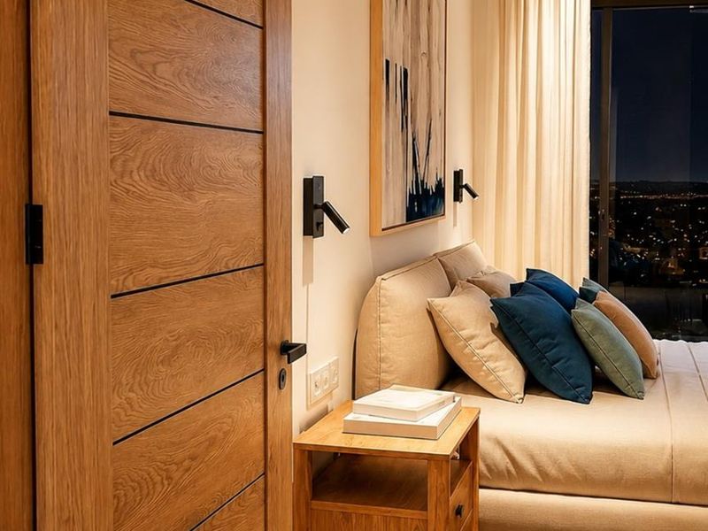 A modern bedroom featuring a sleek wooden door, recessed lighting, and large windows offering expansive city views.