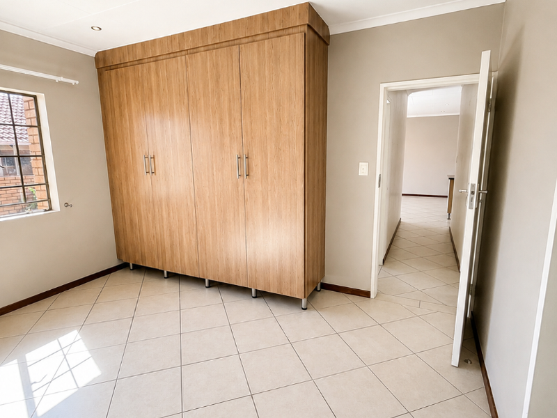 A bright, empty bedroom featuring neutral tiled flooring, built-in wooden cupboards, and recessed ceiling lighting, offering a clean and well-maintained space.