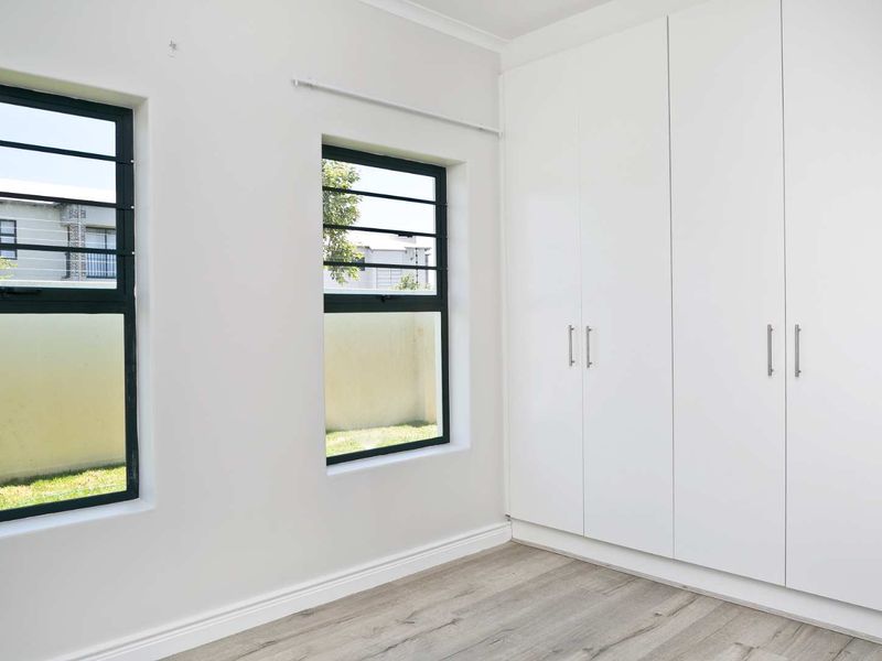 A bright, empty bedroom featuring modern laminate flooring, built-in white cupboards, and aluminium-framed windows with security bars.