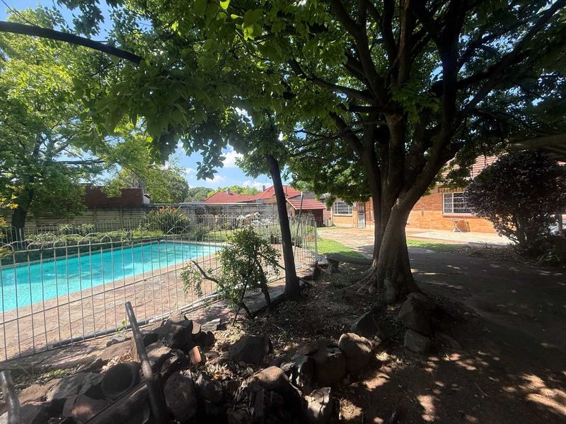 A backyard area featuring a swimming pool with metal fencing, mature trees, and brick paving leading to the main house structure.