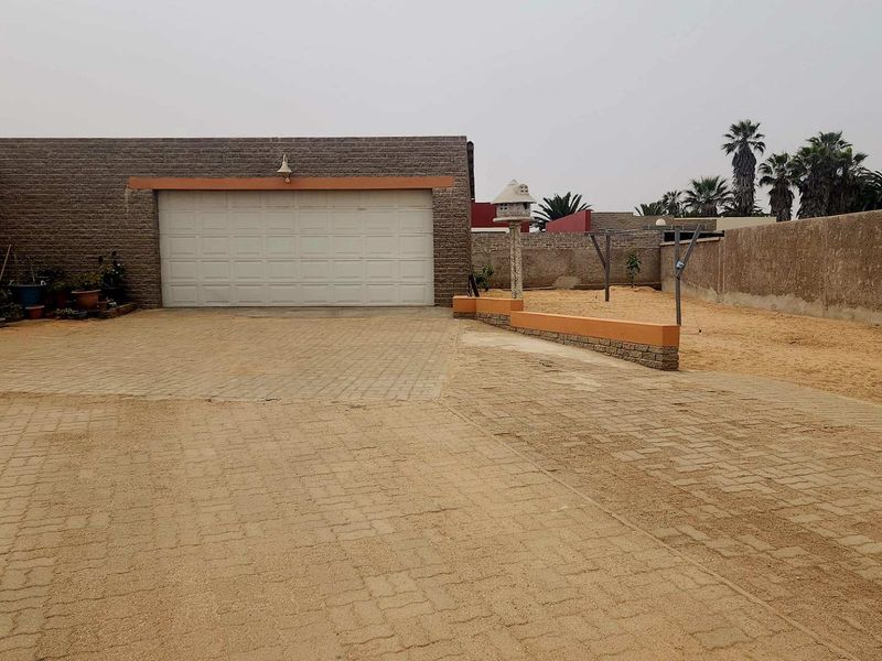 Rear exterior view featuring a paved driveway, a double garage, and a large sandy yard area enclosed by a brick boundary wall.