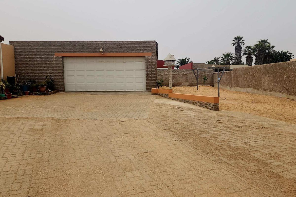 Rear exterior view featuring a paved driveway, a double garage, and a large sandy yard area enclosed by a brick boundary wall.