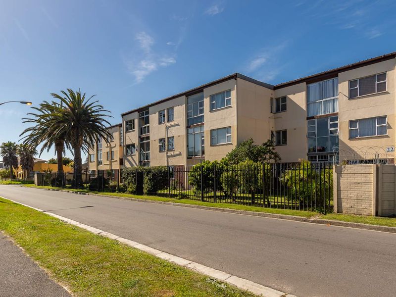 Exterior view of a multi-story apartment building featuring a paved street frontage, metal perimeter fencing, and established landscaping.