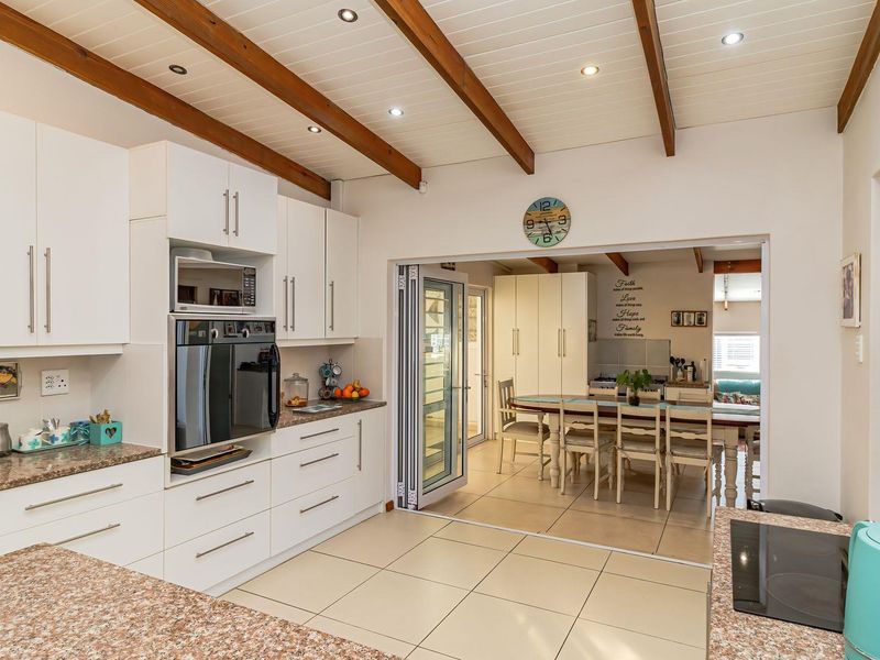 Modern kitchen featuring white cabinetry, granite countertops, built-in appliances, and exposed timber ceiling beams, opening into a bright dining area through bi-fold doors.