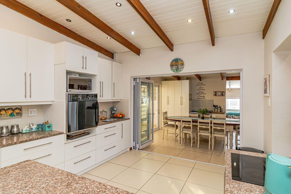 Modern kitchen featuring white cabinetry, granite countertops, built-in appliances, and exposed timber ceiling beams, opening into a bright dining area through bi-fold doors.